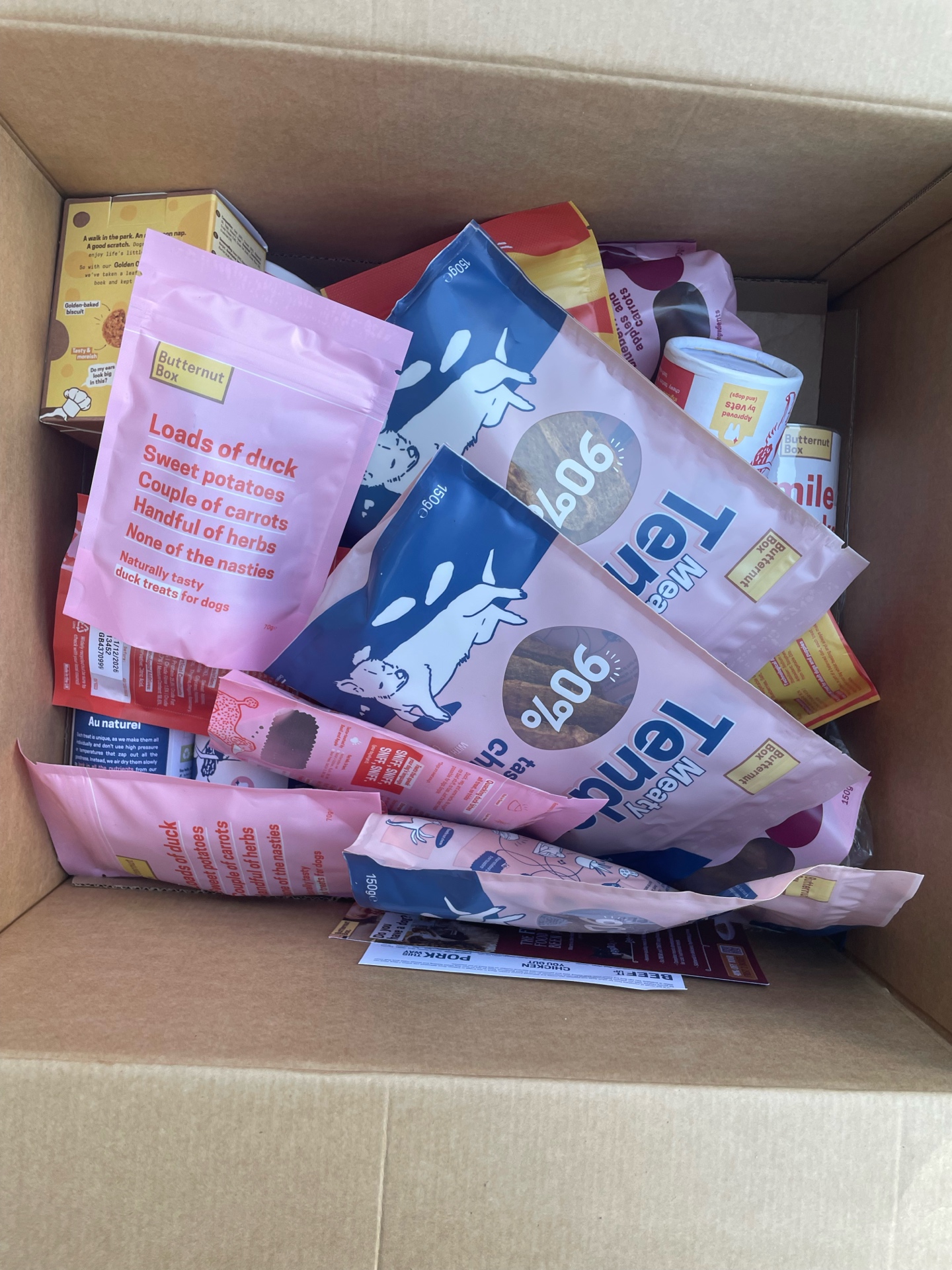 An open Butternut Box showing pink treat bags, blue chicken food bags, cardboard packaging and Tendies.
